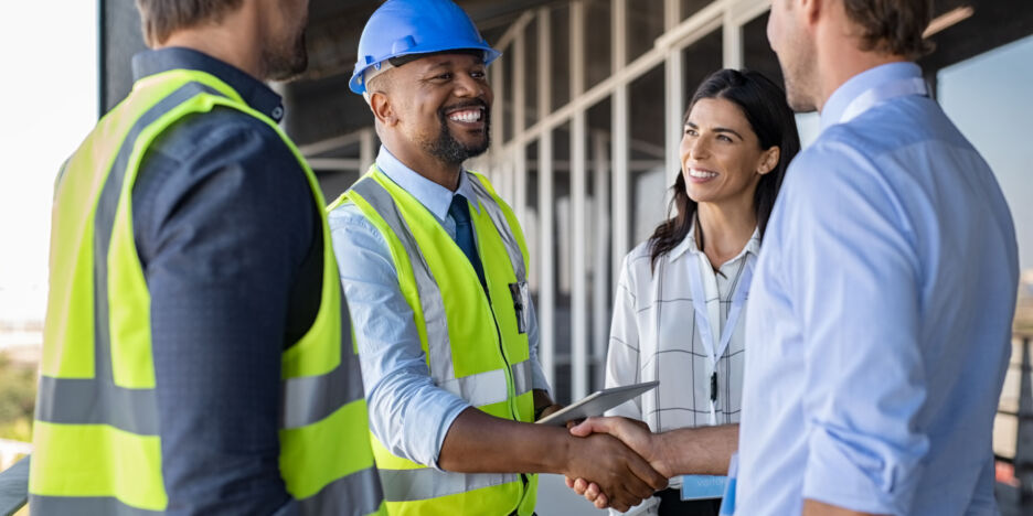 Smiling engineer shaking hands at construction site with happy architect. Handshake between cheerful african construction manager with businessman at bulding site. Team of workers with architects and contractor conclude an agreement with safety uniform.